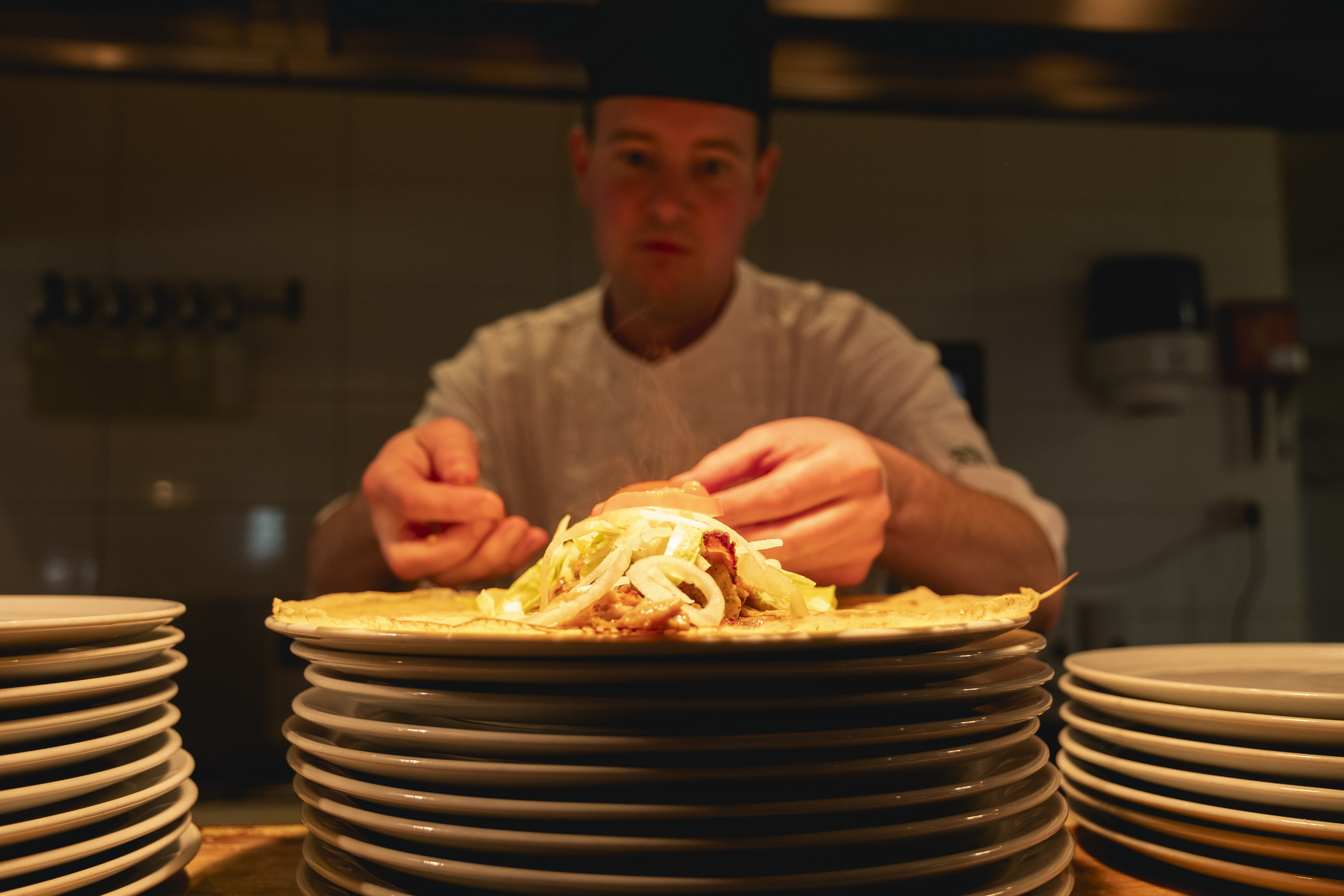Kok houdt een net gevulde pannenkoek boven een stapel borden in de open keuken