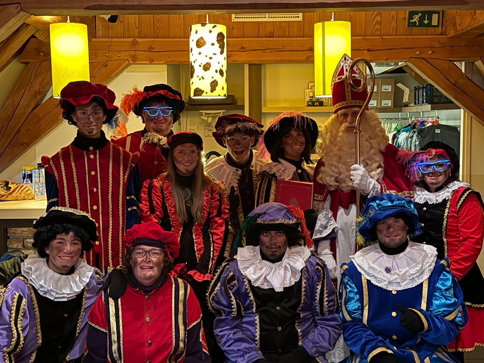 Sinterklaas met mijter en staf en een groep Pieten in kleurige fluwelen kostuums met baretten en witte kragen, samen op de foto onder houten spanten en gele hanglampen
